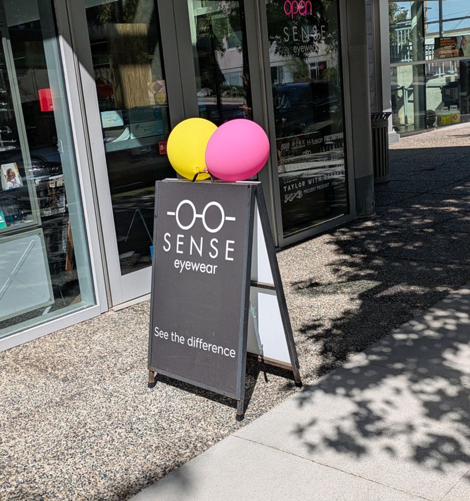 A sandwich board sign for an eyewear store has two balloons on it -- one yellow one pink.