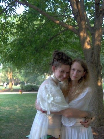 Two teenager girls embrace, cheek to cheek, while looking at the camera.  They both wear white and stand, off-center, in front of a tree.