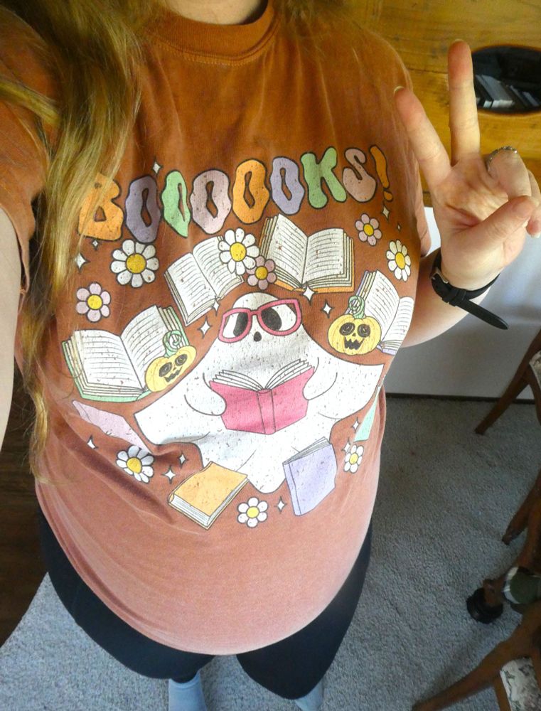 Neck down selfie of a very pregnant person (me) holding up a peace sign and wearing a dusty orange shirt with a picture of a bespectacled ghost surrounded by books and daisies that says in a sort of 70s retro font BOOOOOKS!