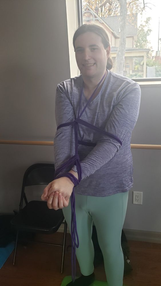 Delia in a purple shirt and teal yoga pants.  Her arms are loosely tied in front of her at her upper arms and wrists