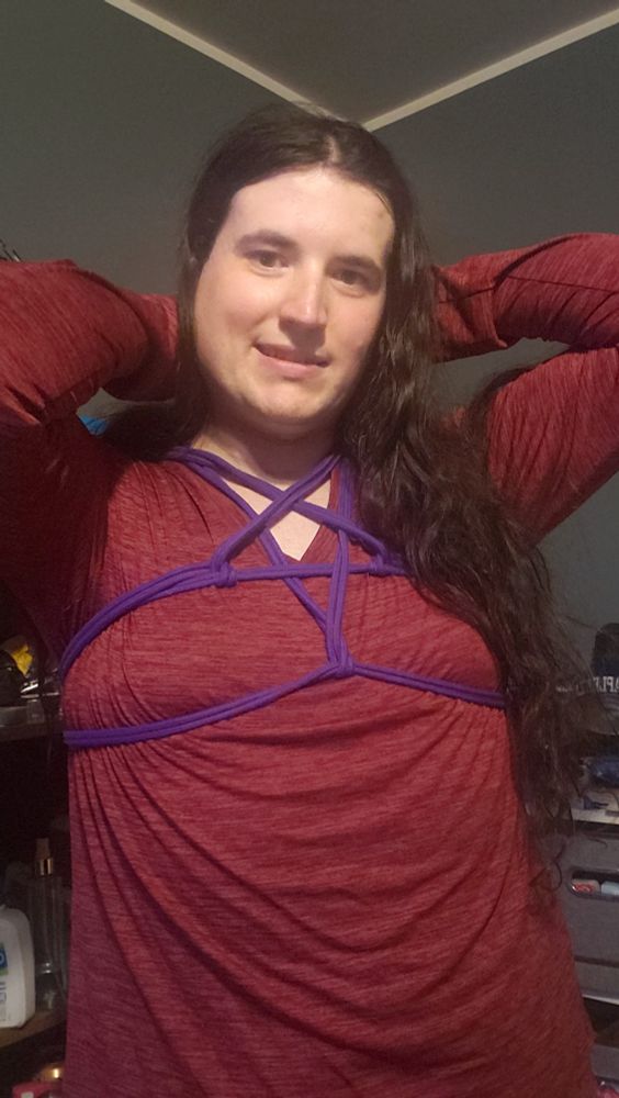Delia in a red shirt with a pentagram chest harness tied with purple rope