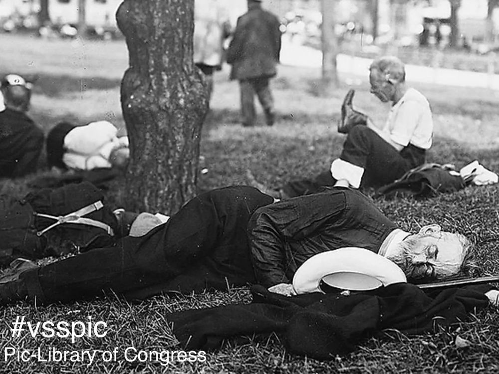 A group of men in old fashioned clothes resting in the shade of a tree. The man in the foreground is lying down, eyes closed. In the back, a man has removed his boot.