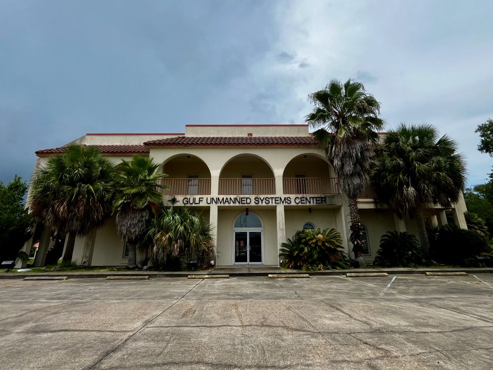A photograph of a tan stucco building with a red tile roof in Carrabelle, Florida. The sign over the door reads. "GULF UNMANNED SYSTEMS CENTER."
