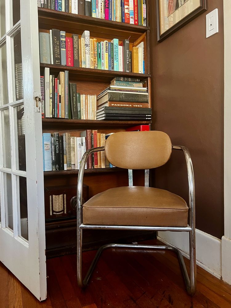 A photograph of a 1940s cantilever chair in front of a bookshelf. The robot vacuum gets hung up on the cantilever part.