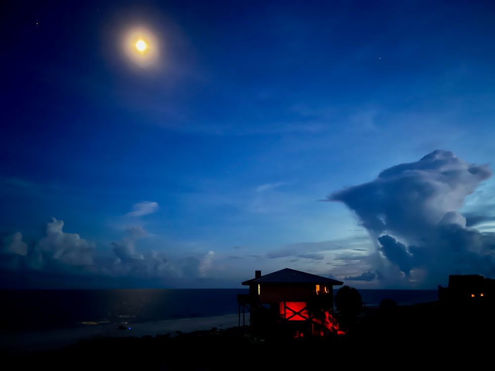A photograph of a house on the beach at St. George Island, Florida. The ocean is in the background. The moon is in the left corner of the photo. A storm cloud is drifting in from the right side of the photo.