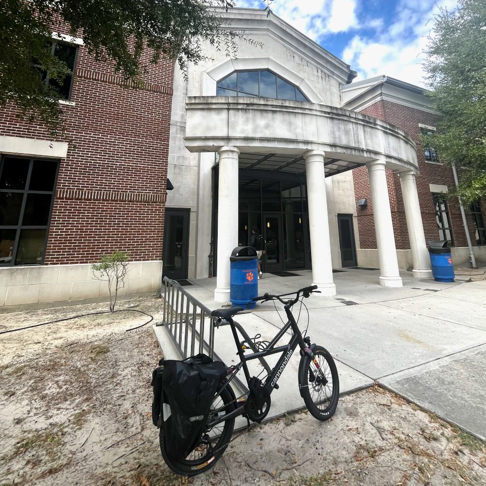 A photograph of a Cannondale Neo Compact electric bicyle locked to a rack outside a campus building in Savannah, Georgia.