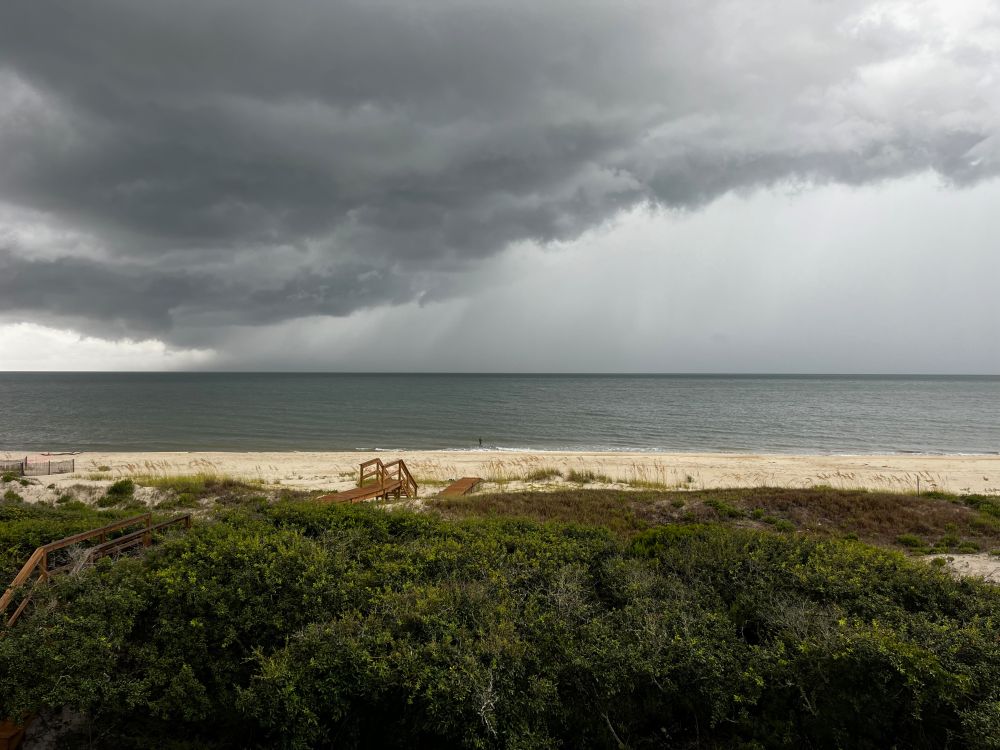 A photograph of the beach at St. George Island, Florida. Dunes and vegetation are in the foreground. A small figure is on the beach fishing. Storm clouds are overhead. 
