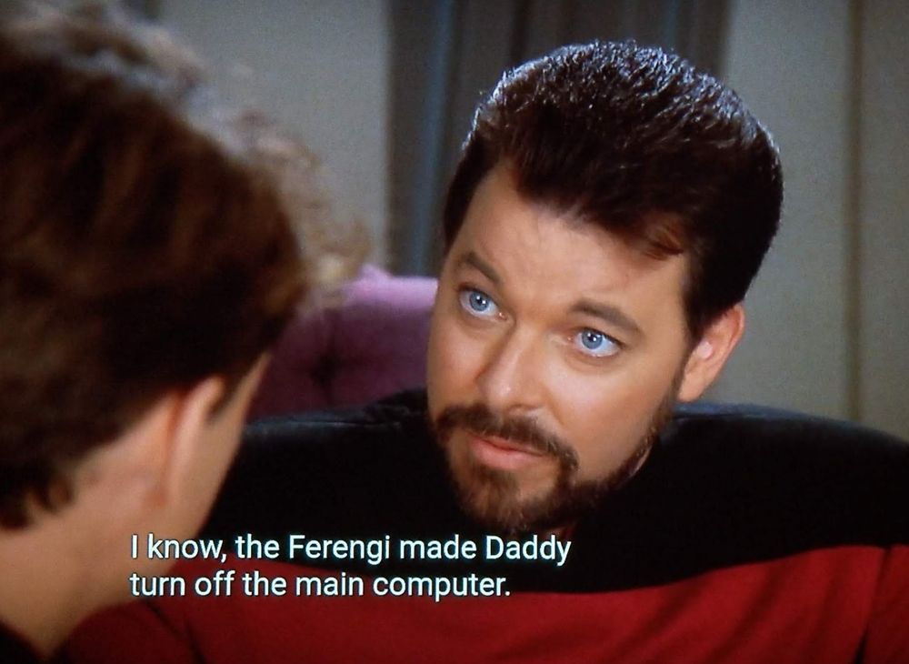 Star Trek The Next Generation scene. We're close in on Riker who is sitting and leaning in and talking to what looks like a youth. His eyes are blue as the ocean and he looks serious in a non threatening kind of way. Closed caption reads, "I know, the Ferengi made Daddy turn off the main computer."