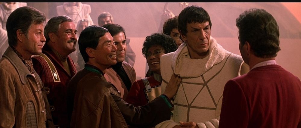 Star Trek The Search for Spock scene. Our Lord and Savior Spock is pictured in a white robe. He stands surrounded by his shipmates who are all older here than they were in the TV show. Sulu is touching Spock's chest for some reason and he's smiling. Uhura, Chekov, Scotty, and Bones also all seem thrilled to see him. Despite being surrounded by friends, Spock is staring only at Kirk, for obvious reasons. 