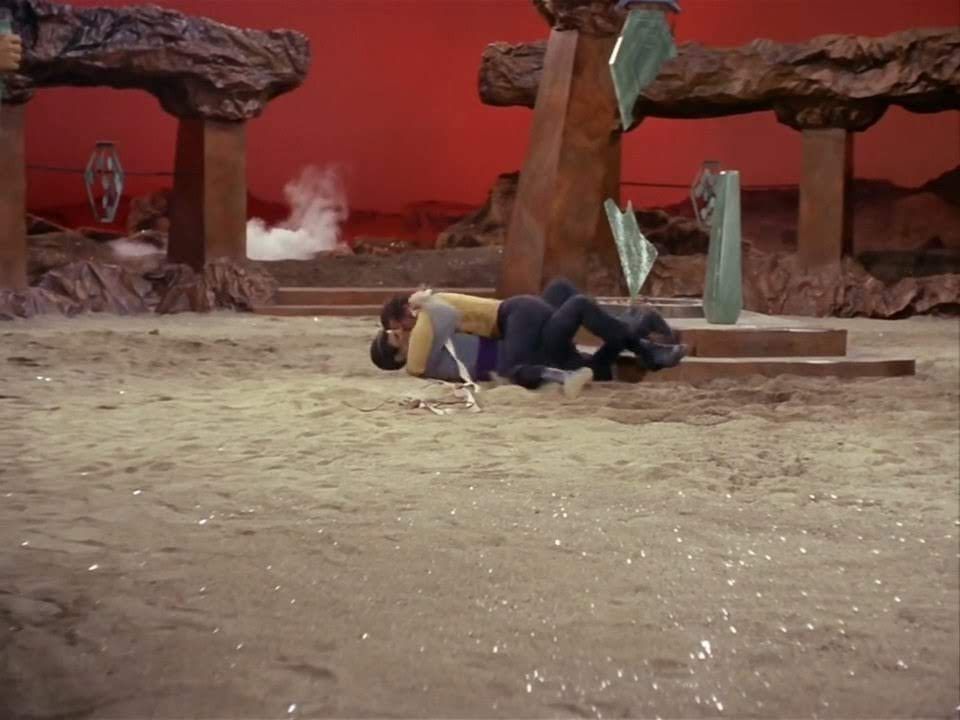 TOS scene. We're outdoors in an arena of some kind with old rocky columns and artifacts surrounding an open dirt area designed for some kind of event or show. A sky colored red lends itself to the passion on display. In the middle, Spock and Kirk are laying on the ground together, Kirk on top, Spock on the bottom, thier bodies intertwined. A love that dareth not speaketh it's name. No caption.