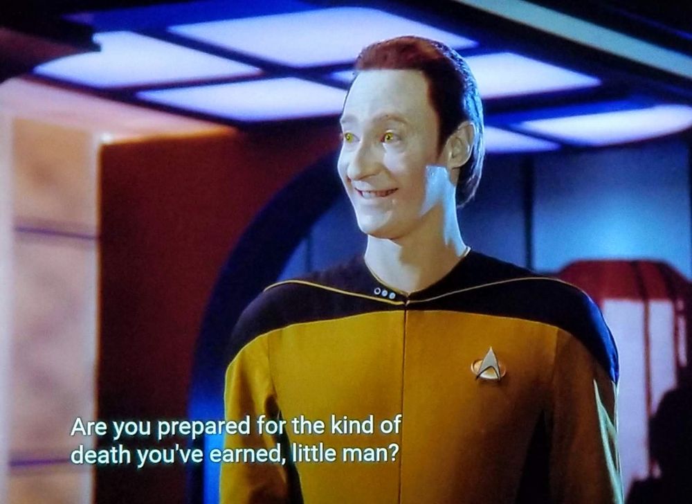 Star Trek The Next Generation scene. We're on ship, possibly in engineering, but it's impossible to know because it's just a screenshot! We're looking at Commander Data, in gold uniform. He's standing and sporting a bright and exaggerated smile. Forehead is very tall, eyes beaming with intense cheer, kind of a horror movie type smile. Closed caption reads, "Are You prepared for the kind of death you've earned, little man?"
