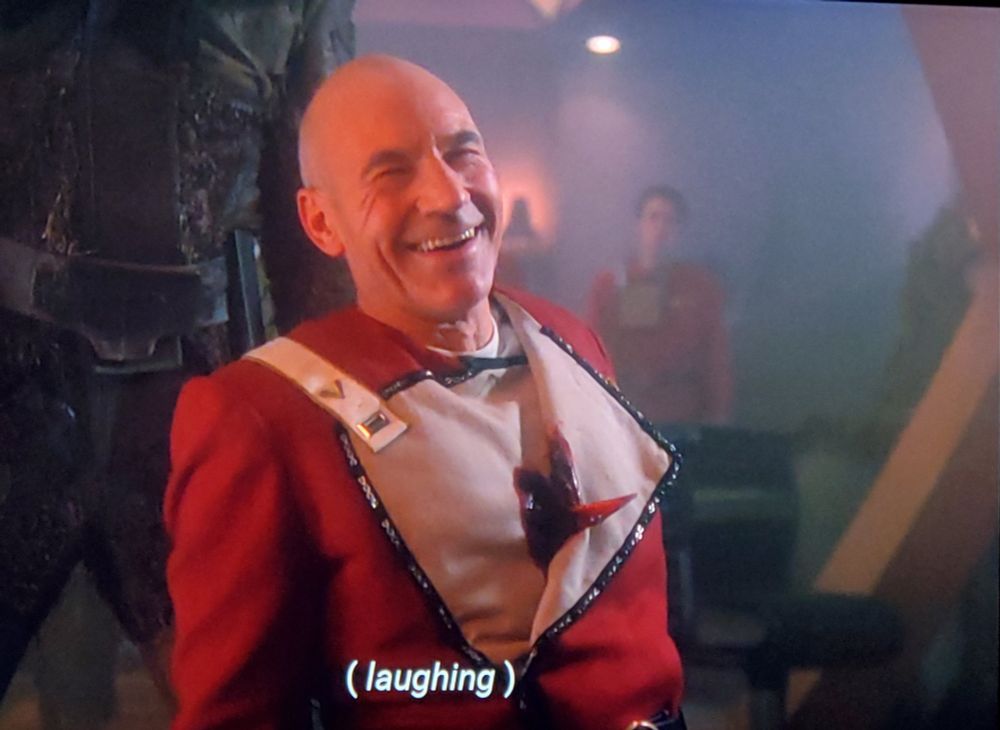 TNG scene. We're in what looks like a smokey room with a starfleet fella in the far background looking on. Picard is sitting down with someone immejiately behind (but you can only see the legs) and Picard is wearing one of the older starfleet uniforms from the TOS films. The "bib" thingy is flapped open and there's a sharp knife protruding out of his chest like someone stabbed him from behind. He's laughing and smiling for some reason. Closed caption reads, "(laughing)"