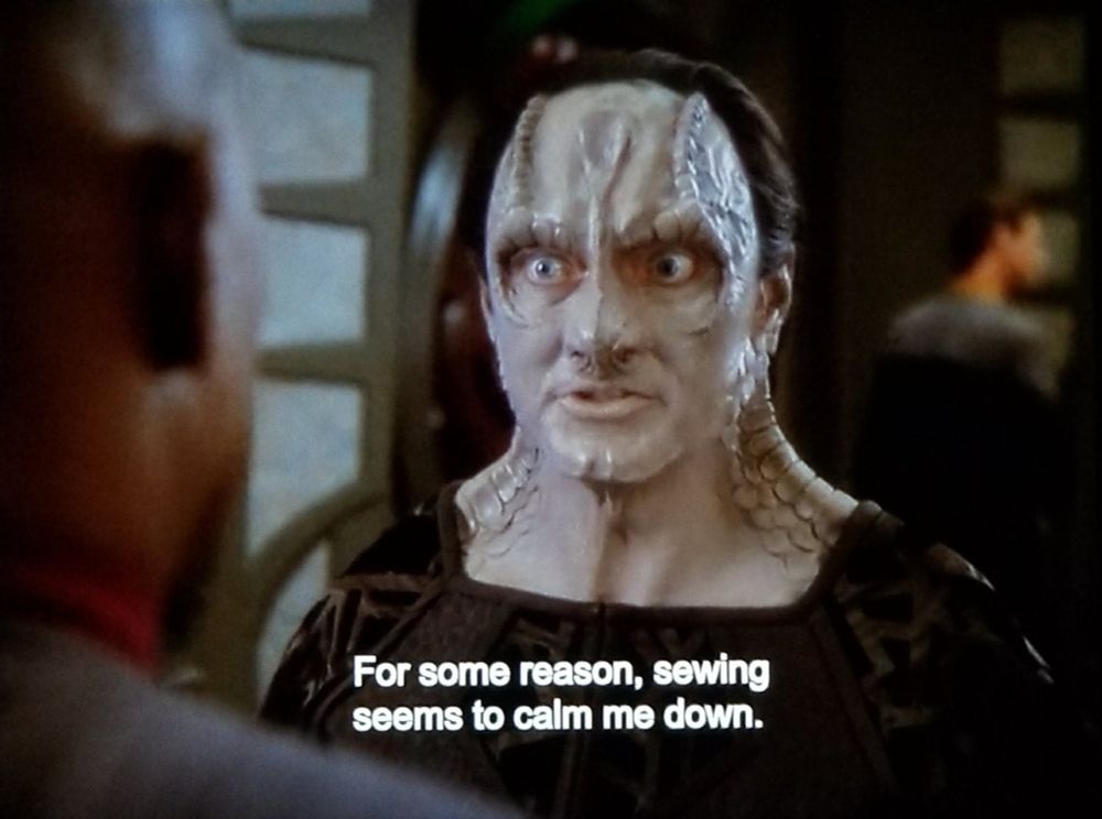 DS9 scene, and it's Garak time. We're on station peeking over the Sisko's shoulder at Garak, a Cardassian with boney ridgey loafy features. He has grey skin and spines that run up his neck, down around his face and eyes, and form a spoon shape on his forehead. He's wearing a low cut get up that highlights an extraordinarily robust neck. Closed caption reads, "For some reason, sewing seems to calm me down."