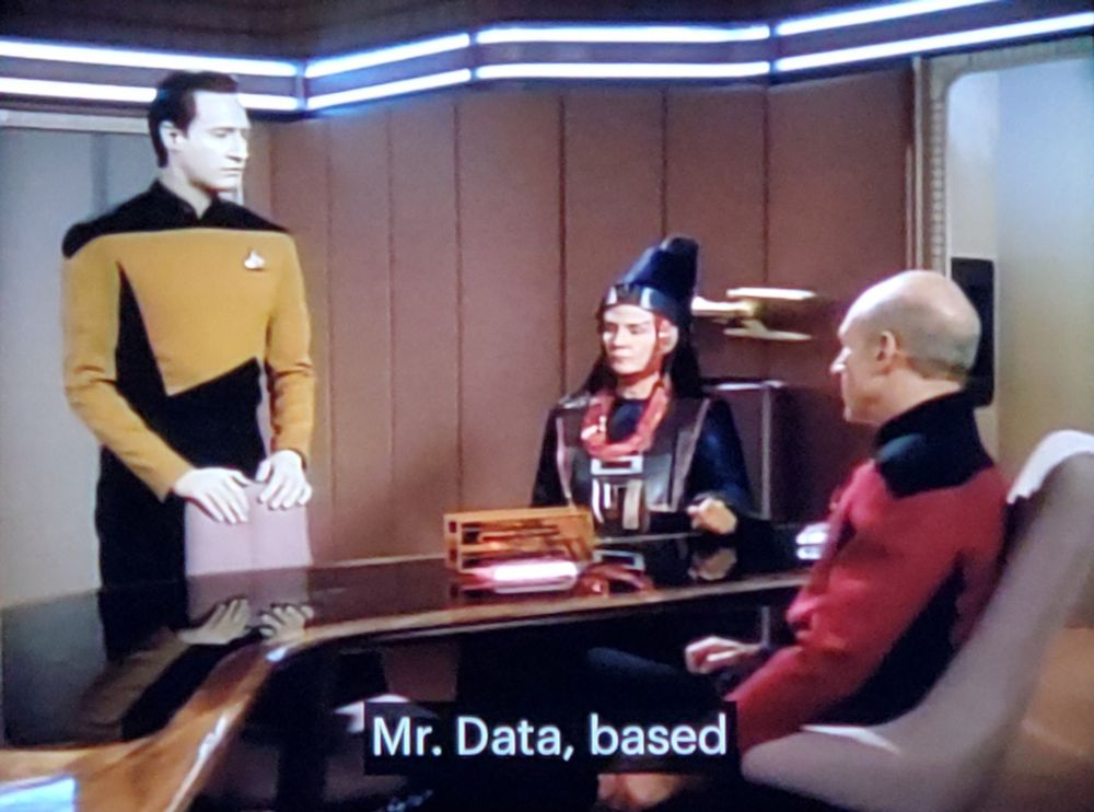 Star Trek The Next Generation scene. We're in Picard's ready room. Picard is sitting like a normal guy at a desk in his uniform and across him is a hoighty toighty looking Vulcan lady with a tall hat and some fancy official looking clothing and accessories sitting across from him. Standing with hands resting on the 3rd chair is Mr. Data, in uniform. 
Closed caption reads, "Mr. Data, based"