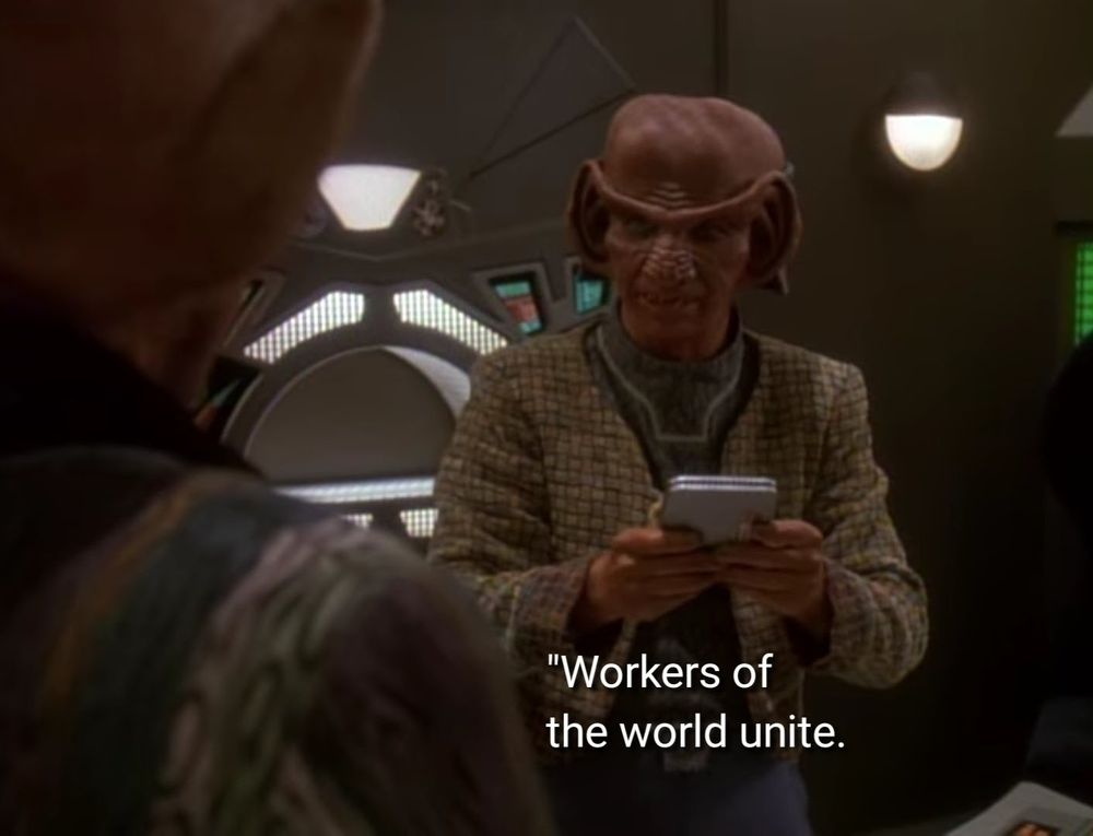 DS9 scene. Rom is a Ferengi hero, with a large bald head and giant ears and lobes that sort of connect to make a unibrow and a ribbed pine cone looking nose. He's wearing a jacket that looks like those lick and stick backsplash tiles you can get at Lowe's. He's pictured here standing in a corridor, talking to someone and holding/reading a PADD. Closed caption reads, "Workers of the world unite. You have nothing to lose but your chains."