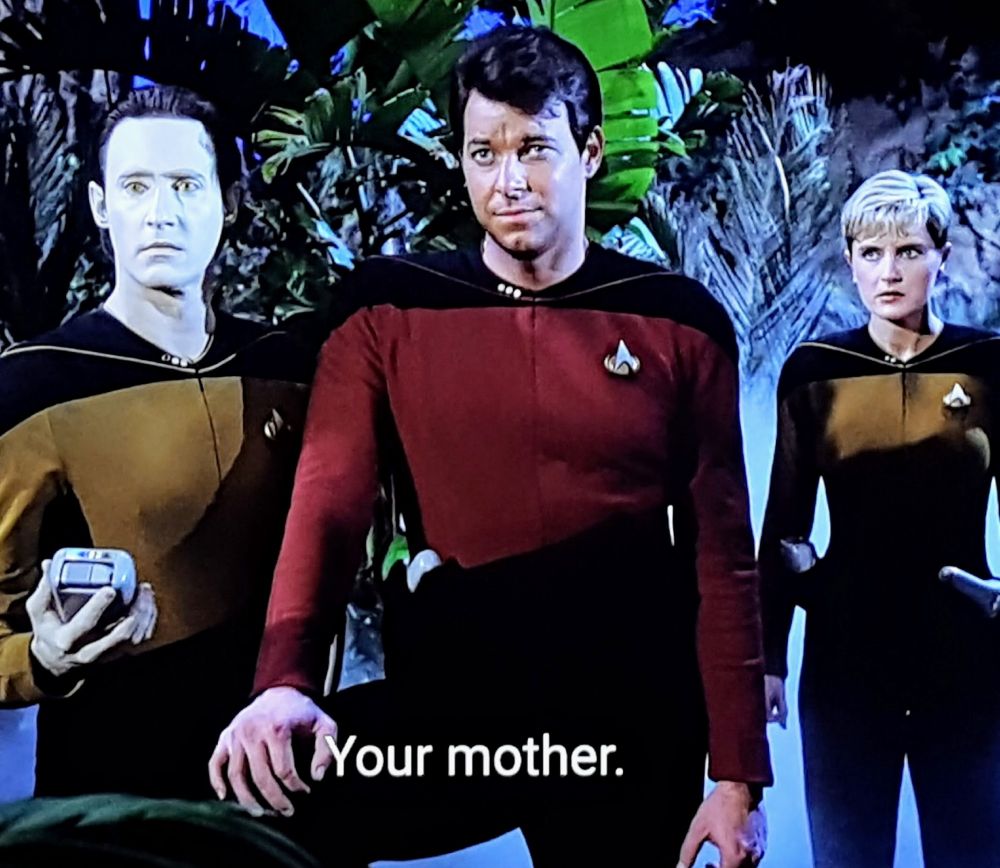 Star Trek The Next Generation scene. We're on a Earthy looking planet with greenery & tropical plants and a foggy haze behind our characters.  Probably quite humid and moist. Centered is Riker, freshly shorn, with Data to his right and Tasha Yar an extra step behind on his left. She looks concerned, Data is pulling a reading on his tricorder, and Riker has his foot propped and his knee up in the classic Riker stance. Closed caption reads, "Your mother."