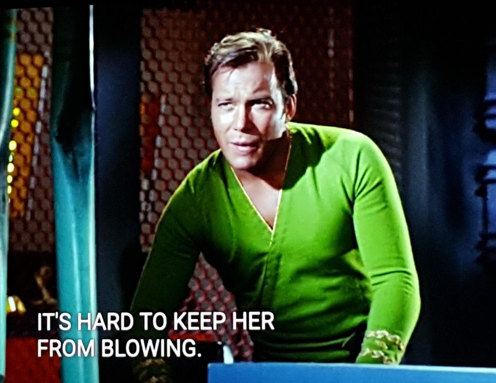 Star Trek The Original Series scene. Kirk is shown on ship and standing at a console with a curious, concentrating look. Kind of holding on like he's on a tippy boat. He's wearing a green uniform with a deep V. Closed caption reads, "It's hard to keep her from blowing."