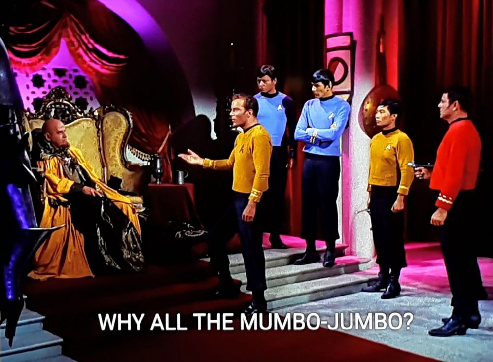 Star Trek The Original Series scene. Ok, so it's a shot of a throne / receiving type room with a lot of pink, purple, and red backlighting and drapery. Sitting on a fancy and elaborate chair with a high back is a bald guy in black clothes and a yellow cape. He seems to the focus of the room. Kirk is standing and addressing him on the steps leading up to him. And behind Kirk is Spock & Bones watching casually, Sulu looking frozen at attention, and Scotty pointing a phaser at no one in particular. There is also like a giant soup ladle hanging as decore for some reason. Ok, I think that's all. Closed caption reads, "WHY ALL THE MUMBO-JUMBO?"