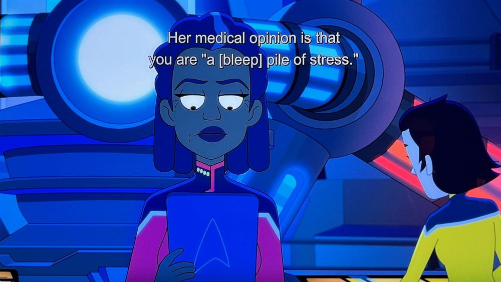 Star Trek Lower Decks scene. Captain Freeman is pictured in Engineering near the warp core which glows blue and red behind her. There's another crew member working in the background. With her back to the previously mentioned warp core, Freeman stands looking down & reading a padd she's holding. Closed caption reads, "Her medical opinion is that you are "a [bleep] pile of stress."