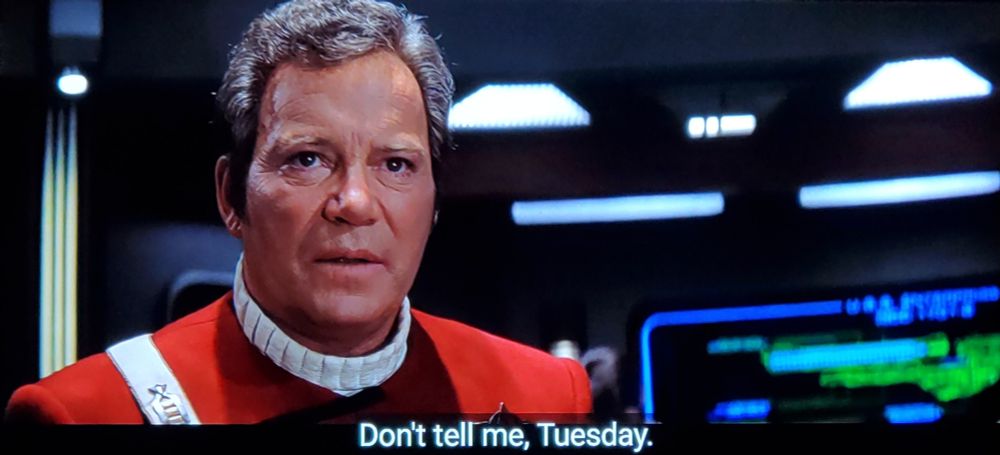 Star Trek Generations scene. Kirk is on the bridge with technical panels and work stations behind him. He's in the classic red movie series uniform with the white marshmallow looking neckline and chest buckle. Here, he is standing and looks concerned.  Closed caption reads, "Don't tell me, Tuesday."