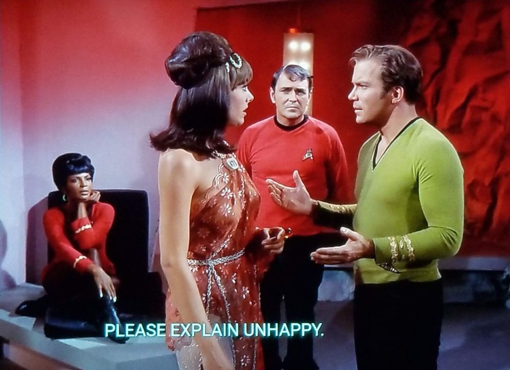 Star Trek The Original Series scene. We're in a red lit away planet room. Kirk is standing and talking to a woman in an up-do haircut wearing a shimmery dress. Scotty is standing behind them looking suspicious. Uhura sitting in the background listening intently. Kirk's hands are extended out in a 'what gives?' motion. Closed caption reads, "Please explain unhappy."