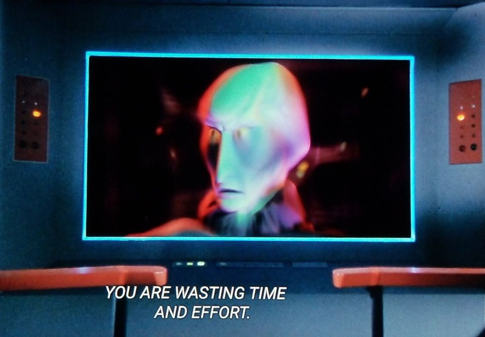 Star Trek The Original Series scene. Enterprise bridge view screen is front and center with an alien head looking off center right. Mr. Alien has a large brain area, long & tall pointed nose, and skin that's greyish with a dash of purple and green. Ominous fellow. Caption READS, "YOU ARE WASTING TIME AND EFFORT. "