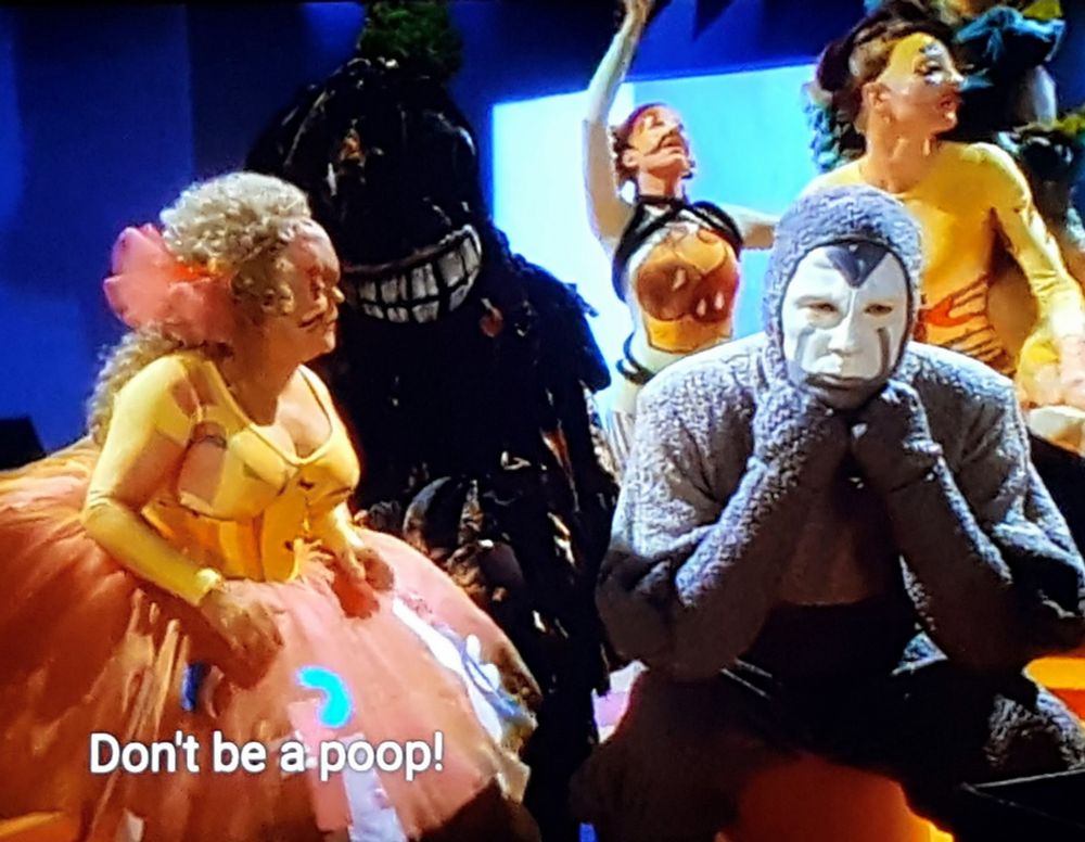 Okay so it's a Star Trek Voyager scene but you can't really tell because we're in some insane horror movie clown room. There's a bunch of weird characters about, like this big round jet black mascot looking furry creature with a giant mouth and menacing white teeth, that thing is standing next to a little person with curly blonde hair in a ballerina tutu thing wearing facepaint. In the background are a couple of ballerina looking carnival folk also wearing Eyes Wide Shut type face masks dancing dramatically. The scary black mascot monster thing and ballerina are focused and looking down towards a sad and sulking clown* wearing a gray outfit that seems made out of a fleece blanket material. (Probably supes cozy) He's sitting like a pouty widdle boy with his elbows on his knees and his chinsey winsey resting on his handsy wandsys. He's wearing gray and white face paint that's drawn like a smile but he's glaring and pissy. Closed caption reads, "Don't be a poop!" 

(* the clown is Michael McKean in real life and he's an American treasure. )