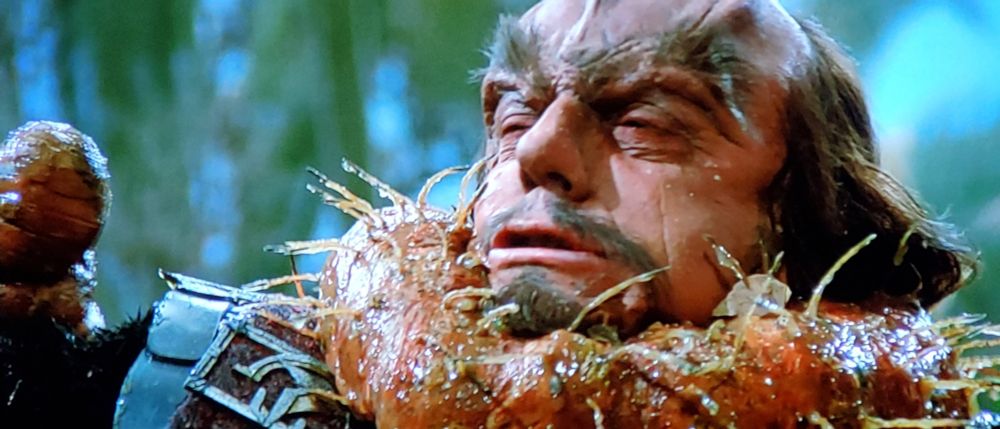 Scene from Star Trek The Search for Spock. It's a series of four pics. In pic 1, a Klingon who looks a lot like the Doc from Back to the Future is standing outdoors on an Earth like planet with trees in the background. He has what appears to be a giant slimy worm with little gross spikey spikes sticking out of it, and the worm thing itself is wrapped around the Klingon's neck and he's holding part of it and it looks painful like he's choking and losing oxygen. Pic 2 is now zoomed out a bit and shows three Klingons, 2 are watching the main Klingon who is struggling with the large gross worm monster, who is no longer being choked, and he is now holding the worm threateningly out in front of him, though it remains draped around him. Pic 3 shows the Klingons continuing to watch as main Doc Klingon is taking the gross worm thing off of himself and it seems now like the risk has been averted. Pic 4 shows shows Doc Emmit Brown Klingon standing by the 2 onlookers, and he's holding a communicator near his face. Closed caption reads, "Bridge, nothing happening here."