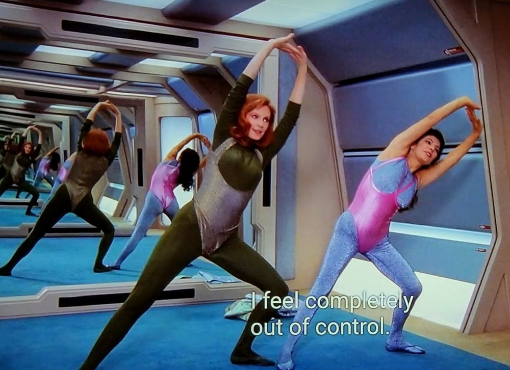Star Trek The Next Generation scene. We're in a carpeted workout room on ship with a mirror back wall that looks oddly & exactly like if they just put a mirror up in the Enterprise D corridor and decided to get their sweat on in the hallway. On screen is Troi and Crusher, wearing rad! jazzersize outfits from the 80s. Like leotard bodysuits with alternating pink and green and silver and glittery colors. Both are standing and leaning and stretching thier arms overhead with smiles. Behind them you see an infinity loop of them working out going into the background of mirror reflections. Closed caption reads, "I feel completely out of control."