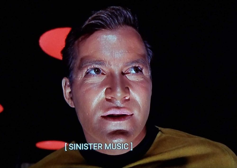Star Trek The Original Series scene. We're uncomfortably close in on Kirk, shot from a little under and looking up at him. We're in the transporter room but the lights are low and red so it's dark and eerie. He's a bit sweaty and moist looking, and has a scheming look on his face. Closed caption reads, "[SINISTER MUSIC]