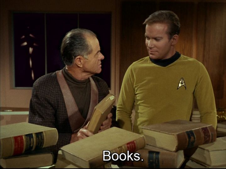 Star Trek The Original Series scene. We're on an away mission somewhere in a room. We see Kirk in uniform and a another not Starfleet fella. The non Starfleet fella is holding a book. In front of them both is a pile of old looking books. Kirk looks weirdly into the old guy. Closed caption reads, "Books."