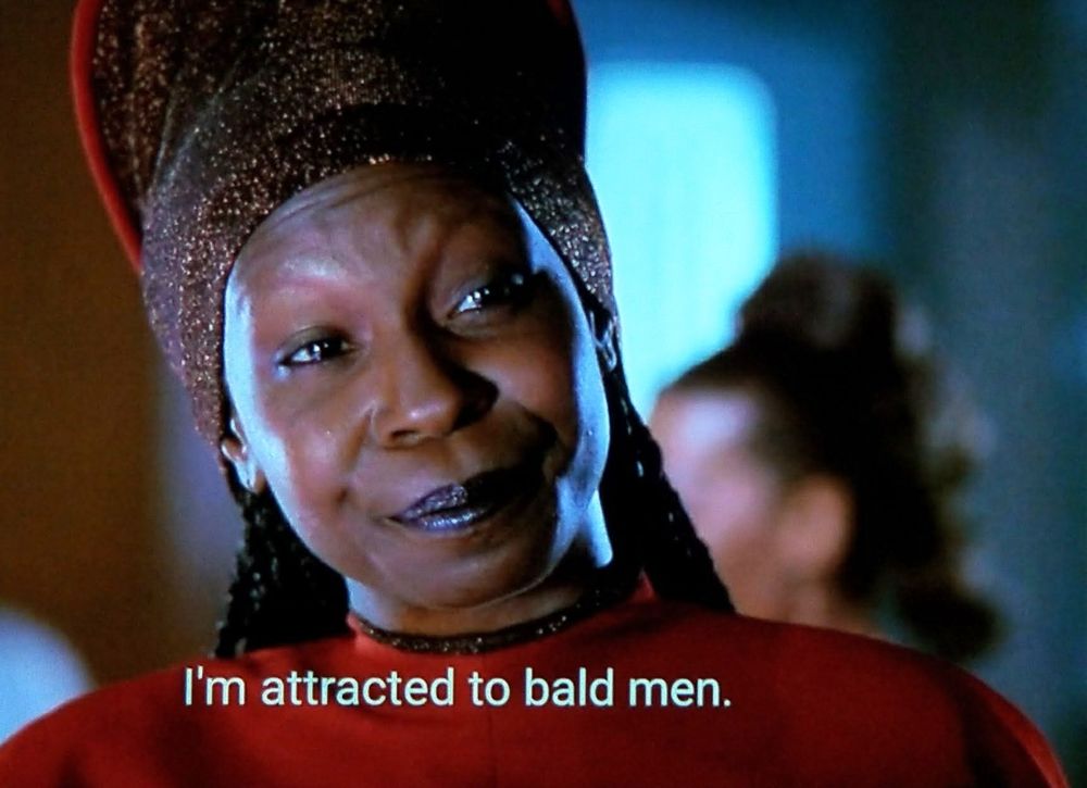 Star Trek The Next Generation scene. We're close in on Guinan, a humanoid looking alien who is the bartender in Ten Forward. She wears tall and cool hats which are rounded or tall and flat and always on point fashion wise. Closed caption reads, "I'm attracted to bald men "