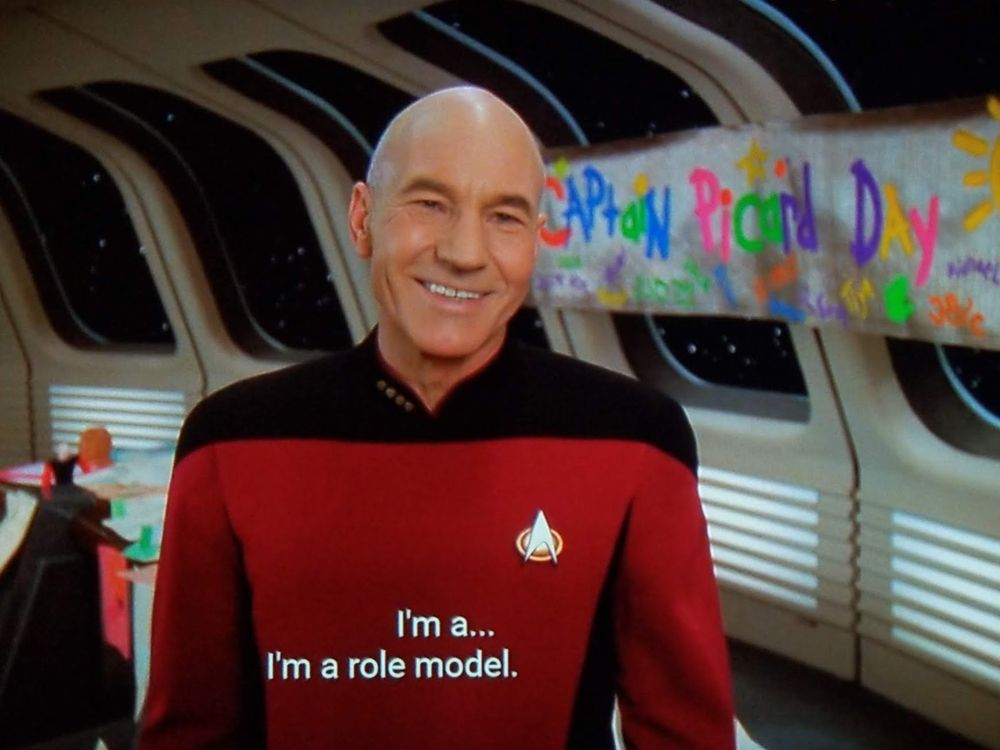 Star Trek The Next Generation scene. We're on the Enterprise, in a room with many windows out to space. It looks like a classroom type setup with a table and crafts in the background, but also probably Ten Forward with a different set. Captain Picard stands and has a big awkward smile on his face. Behind him, a banner is hung with 'Captain Picard Day' drawn on it. The letters are different colors and clearly drawn by children, like if you were a righty but wrote with your left. The banner also has different cute little other drawings around the letters, like flowers, hand prints, and a sun. Closed caption reads, "I'm a...  I'm a role model."