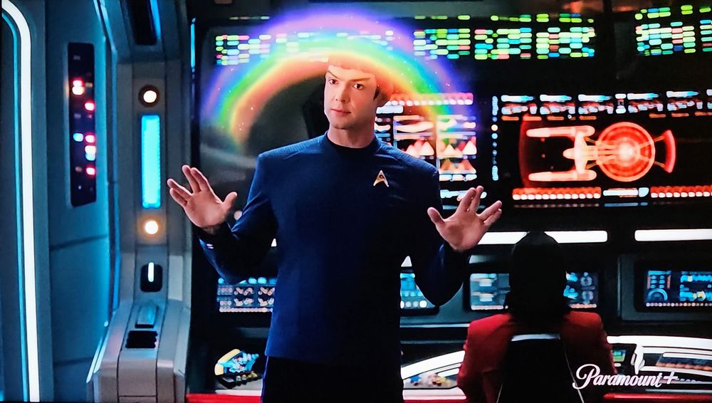 It's an image from a promo for Star Trek Strange New Worlds. Spock, in blue uniform, is standing on the bridge with a full array of consoles and touchscreens behind him for bleep bloop ship functions. He's making the live long and prosper sign with both hands and a colorful rainbow arches between them. 