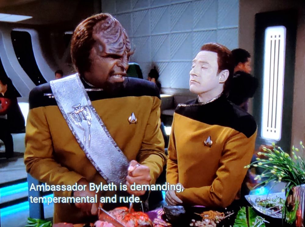 Star Trek The Next Generation scene. Ok, so it's four screenshots and we're having a fancy dinner party in Ten Forward. There's guests sitting in the background and we're focused on Worf & Data. Worf's #BattleSash is extra fancy that's how you know we're at a party. Worf is carving what looks like a turkey on a table with lots of different foods and flowers on it, and looks VERY irritated while Data is speaking. Closed caption reads, "Ambassador Byleth is demanding, temperamental, and rude. You share all those qualities in abundance. Perhaps you should try to build on your similarities." In the third screenshot Data gives Worf a patronizing shoulder pat. 
The fourth screenshot shows Worf holding up the very large carving knife towards Data who has just walked away. Worf looks like he wants to shank him. 
