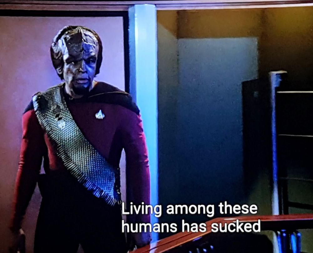 Star Trek The Next Generation scene. We're in a darkly lit engineering near the warp core. Worf is in uniform and walking alongside the cute lil safety railing. He looks focused and intense. Closed caption reads, "Living among these humans has sucked."