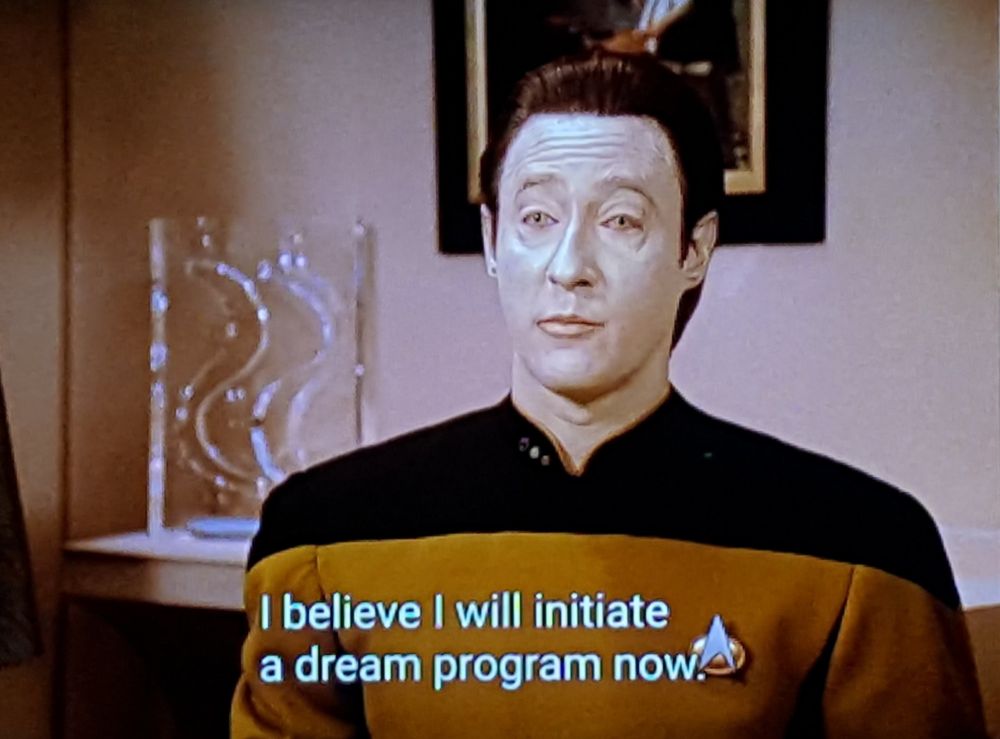 Star Trek The Next Generation scene. Here we see Mr. Data, in quarters, with a flowey fancy glassy art piece behind him on a shelf. He's just standing and talking otherwise, in his nice golden uniform. Closed caption, "I believe I will initiate a dream program now."