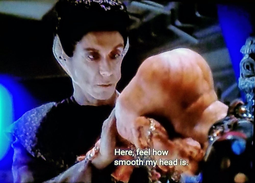Star Trek Deep Space Nine scene. Ok, so Iggy Pop is a Vorta which is a humanoid alien with tall swooping up ears along the edge of the face, super pale skin, and jet black hair. Here, he is standing and touching the large bald head of a bowing Ferengi who as we know have heads that kind of look like butts with large and round lobes almost equal to the size of their heads. Closed caption reads, "Here, feel how smooth my head is."