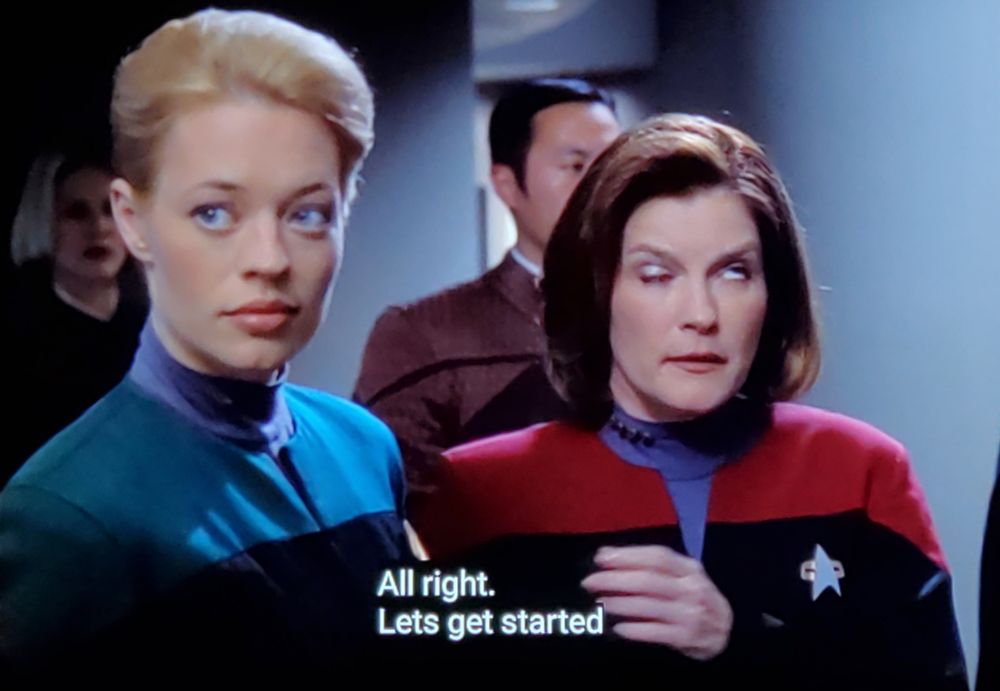 Star Trek Voyager scene. We're IN TRANSIT in a corridor walking with Captain Janeway and Seven on Nine (Tertiary adjunct of unimatrix 01). They're both looking official in their red and blue uniforms and there's some other folks/extras behind them also walking. Janeway has one eye open looking straight up and one eye closed in pic 1, and in pic 2 looks like she has no time for anyone's bullshit whatsoever. Closed caption reads, "All right. Let's get started before my headache gets any worse."