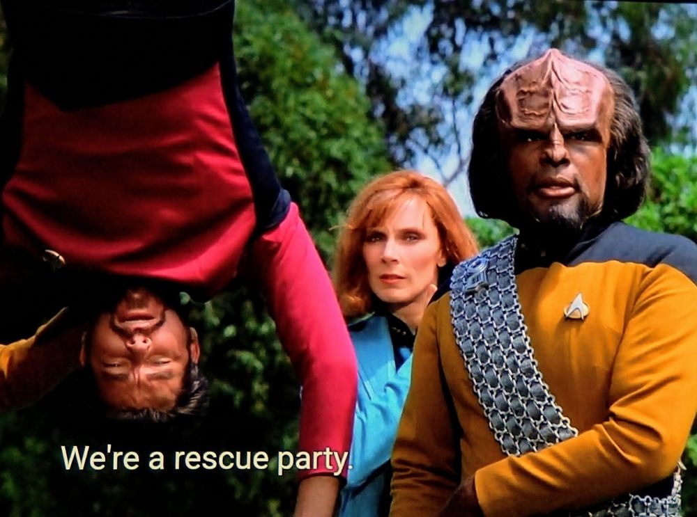 A bright and beautiful Next Generation scene here. We're outdoors with green trees in the background. The shot is centered on Worf, Crusher, and Riker. Worf in gold, Crusher in blue, and Riker in red. Riker is hanging upside down with arms danglin'. Closed caption reads, "We're a rescue party."