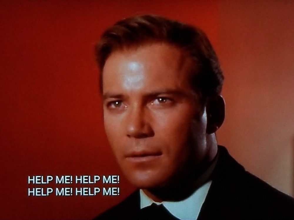 Star Trek The Original Series scene. Kirk's shiney younger actor face is shown, close up. Looks like  he's wearing a suite and tie, which is kind of weird since he's a future space guy. The background is a gradient of red to less red. He looks like he's listening to something vaguely interesting. Closed caption reads, "HELP ME! HELP ME! HELP ME! HELP ME!"