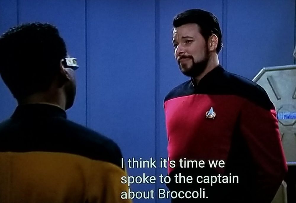 Star Trek The Next Generation scene. We're in the CARGO BAY! Riker in red uniform is speaking with Geordi in gold uniform. Riker has a smug sort of skeptical look on his face as he listens. Closed caption reads, "I think it's time we spoke to the captain about Broccoli."