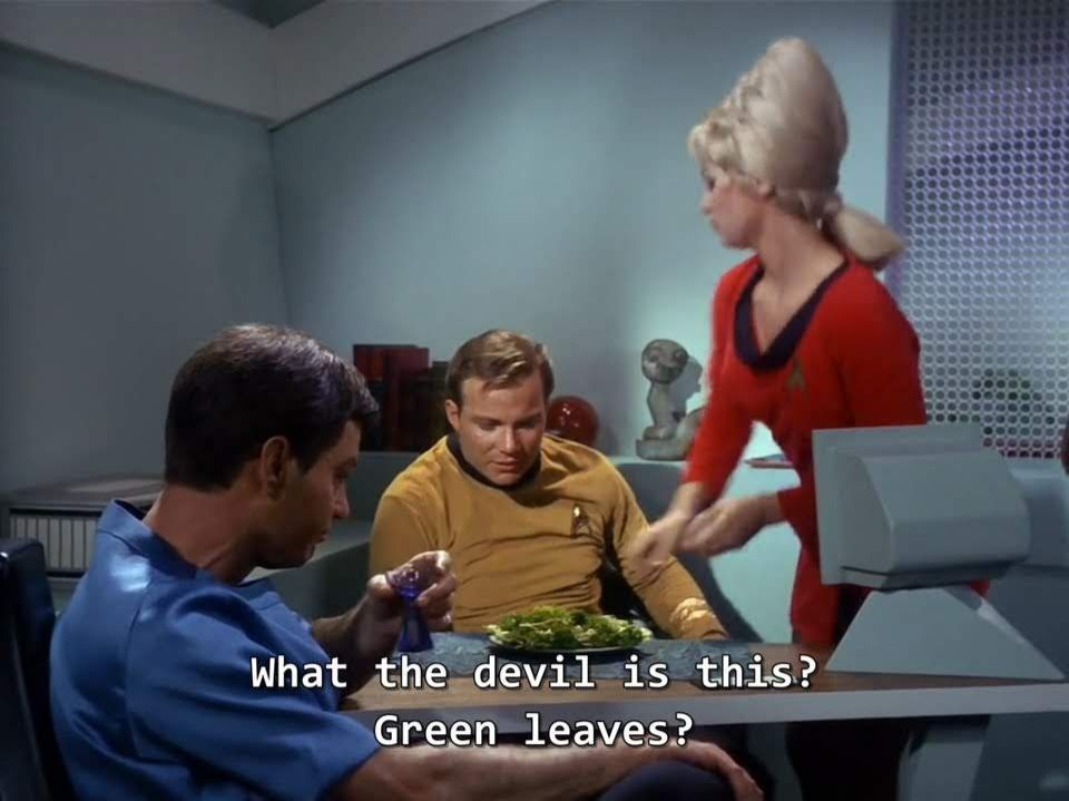 TOS scene. We're on the Enterprise I think in Kirk's quarters. Sitting is Dr McCoy and Kirk in uniform. McCoy is holding a shot glass looking drink. Standing near is Yeoman Rand who looks like she just dropped off a salad for Kirk. Kirk is sort of taking it all in visually. Closed caption reads, "What the devil is this? Green leaves?"