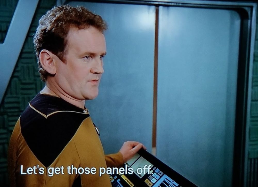 TNG scene. Chief O'Brien, who can do a wheelie on a unicycle, is standing in the transporter room. Chief O'Brien, who can slam a revolving door, is looking off to the right. Chief O'Brien, who can build a snowman out of rain, is wearing his standard gold uniform. Closed caption reads, "Let's get those panels off."