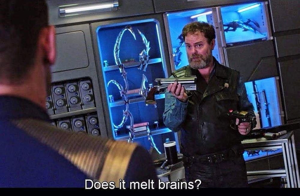 Star Trek Discovery scene. Dwight from the Office, but here as a Star Trek guy, is standing and holding up some machine part in one hand and a phaser in the other. He's dressed in non civilian bad boy rogue type clothes and the room is busy with other techy stuff and weapons and tubes and tables. Closed caption reads, "Does it melt brains?"
