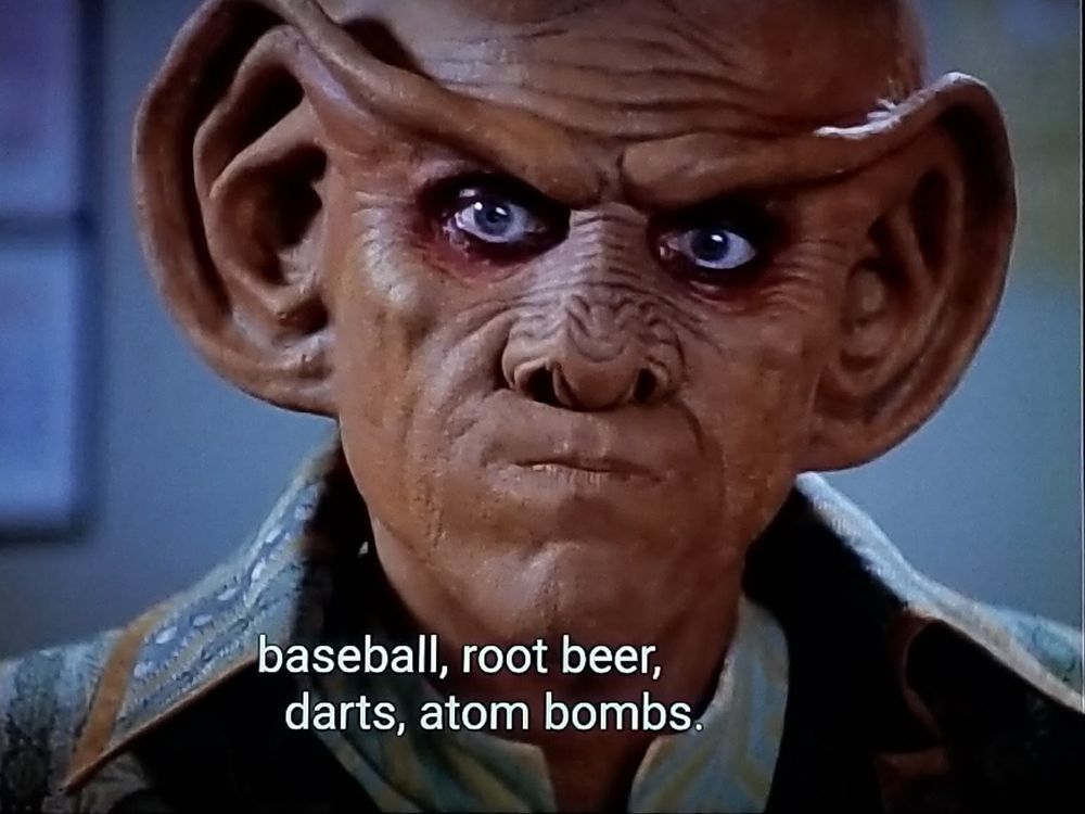 Star Trek Deep Space Nine scene. Close up on Quark, a Ferengi with a smooth and domed head and huge earlobes that connect like a unibrow across his face. He also has nose loaf that kind of looks like a pine cone.  He is pursing his mouth and doing a Jim from the office look right at us. Closed caption reads, "baseball, root beer, darts, atom bombs."