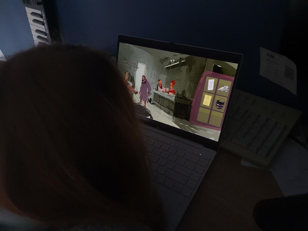 Back of the head of a long haired child, looking at a computer screen showing a grey interior of a inn, with several characters.