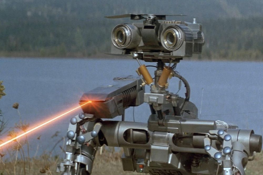 Johnny Five from the movie Short Circuit standing in front of a body of water and shooting a laser beam out of his shoulder thing