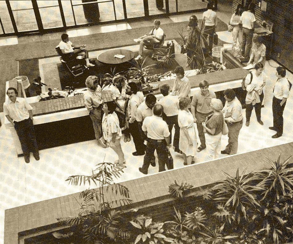 A black and white photo, taken from above, shows the atrium of the College of Social Work, with small plants and a couple dozen people talking to each other