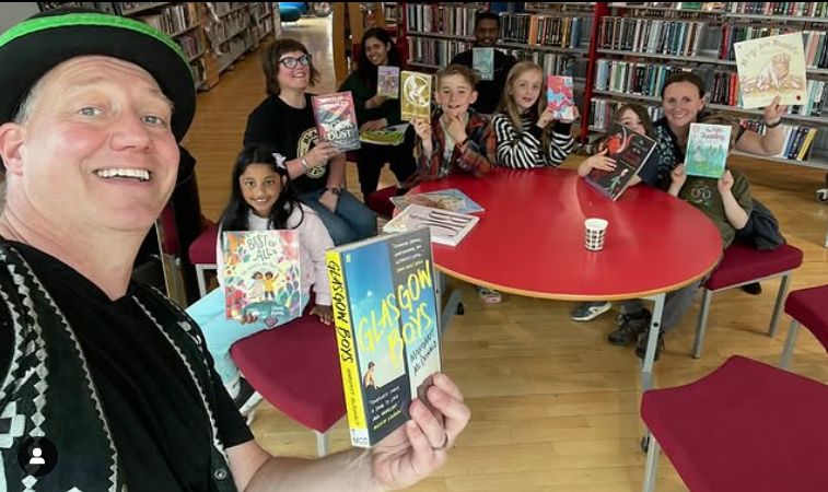 Poet Paul holds a copy of Mary MacDonald's Glasgow boys families sit smile at the camera from around a round table in the background.